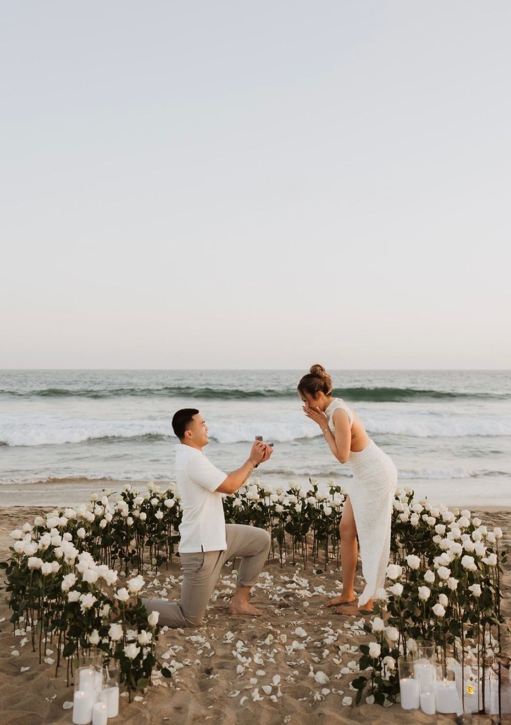 A proposal inside a circle of white roses and candles on the beach at sunset